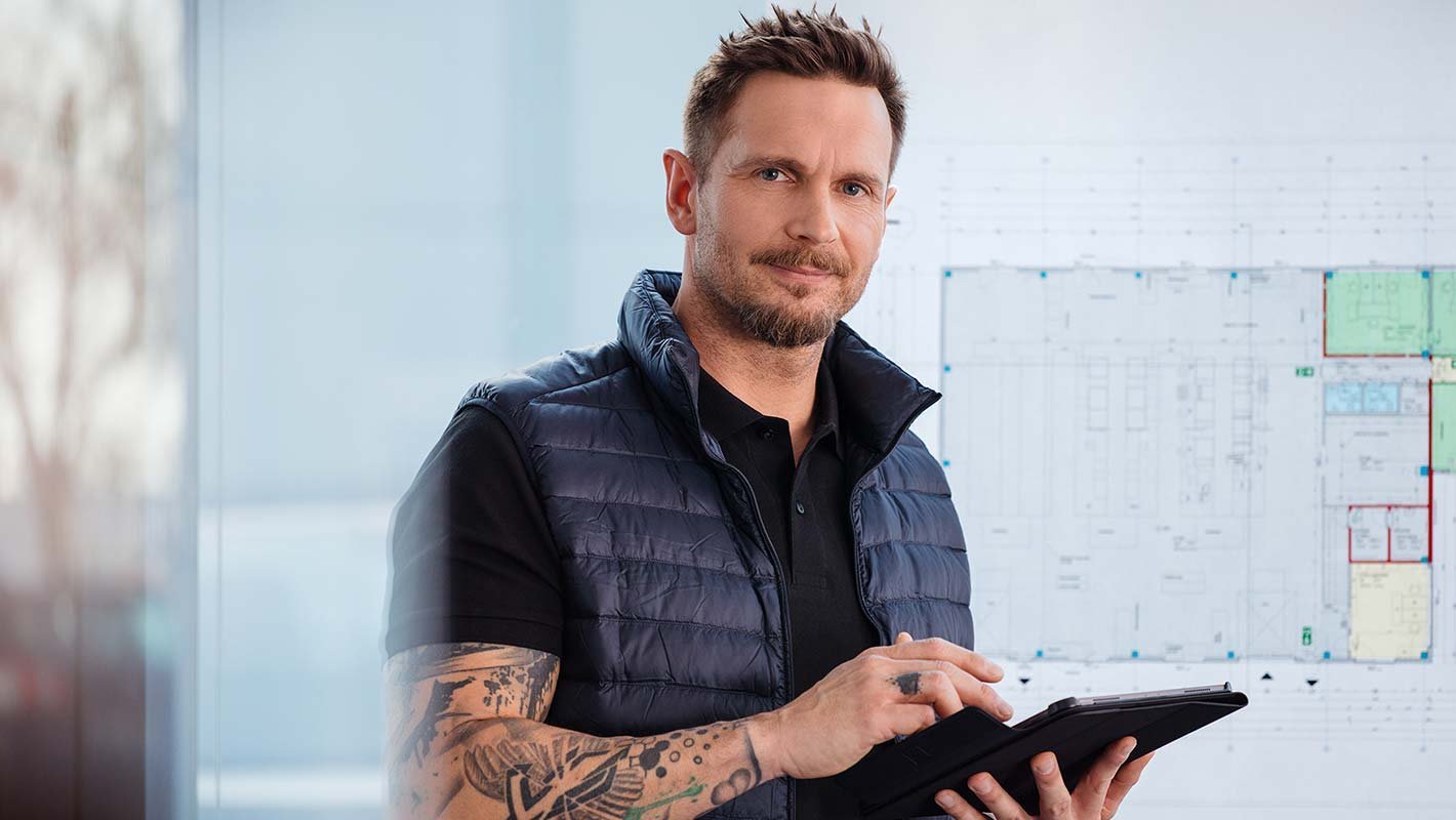 Ein Mann hält ein Tablet in der Hand und im Hintergrund ist eine Tafel an der eine Technische Zeichnung hängt