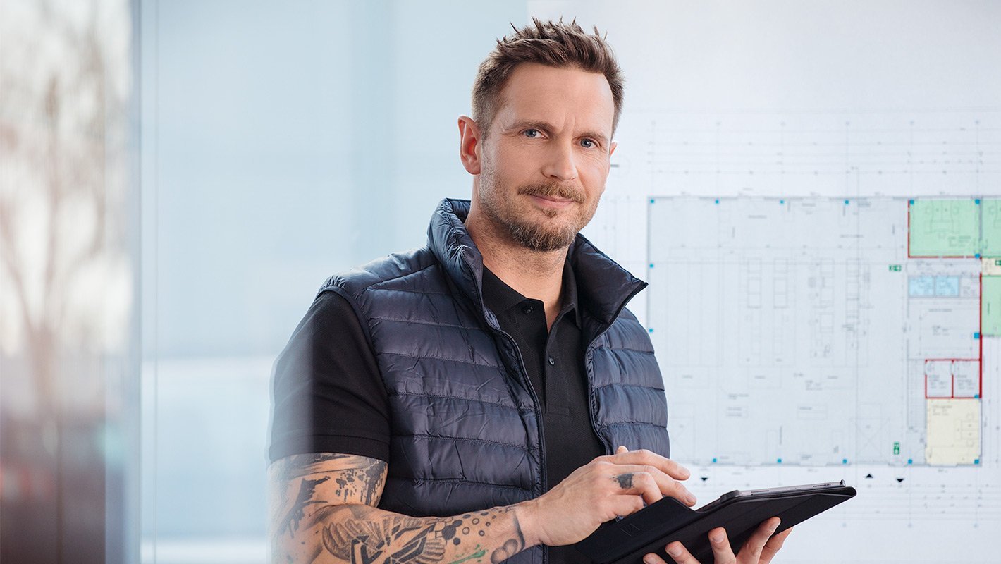 Ein Mann hält ein Tablet in der Hand und im Hintergrund ist eine Tafel an der eine Technische Zeichnung hängt
